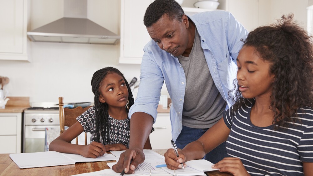 Father Helping Two Daughters Sitting At Table Doing Homework